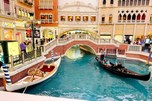 Views of a blue lagoon waterway with an arched bridge, and two gondola boats, one with passengers, at a Venice-style waterfront village at the Venetian Resort in Vegas.