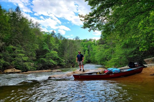 A hiker standing next to a canoe on the banks of a scenic river in Dawsonville, GA, surrounded by a heavily wooded area.