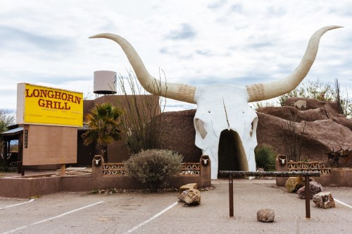 The entrance of a roadside attraction in Arizona featuring a massive skull with 30-foot-tall longhorns tucked against a rock building structure with a large yellow sign and several cars parked outside.