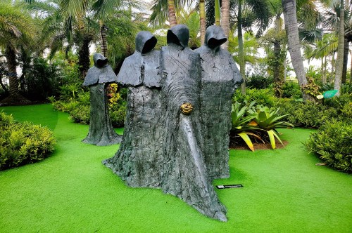 A sculpture featuring four mysterious hooded figures surrounded by tropical flowers and palm trees, inside a sculpture garden in Orlando, FL