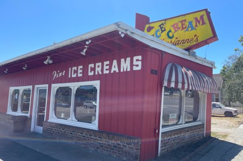A small red building with white details and lettering saying, 'fine ICE CREAMS,' with an old-fashioned yellow sign above saying, 'Marianne's Ice Cream.'