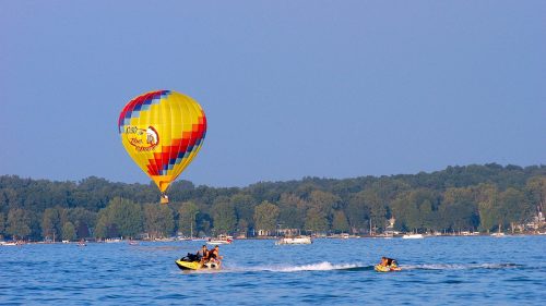 A colorful hot air balloon rises above a the blue water of a lake on which a jet ski tows a tube, while a pontoon and other watercraft ply the waters in the background.