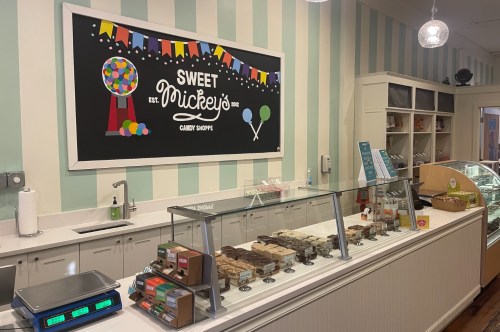 The inside of a candy shop, the front desk has fudge displays and the back wall has a sign that reads, 'Sweet Mickey's Candy Shoppe.'