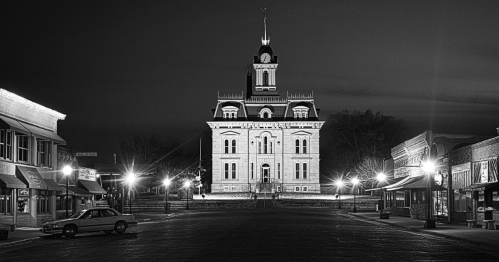 A black and white night photo of a historic stone courthouse with a clock tower, centered on a quiet, empty street.
