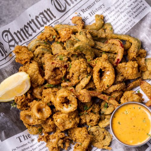 A pile of crispy fried seafood and vegetables on newspaper, served with a lemon wedge and dipping sauce.