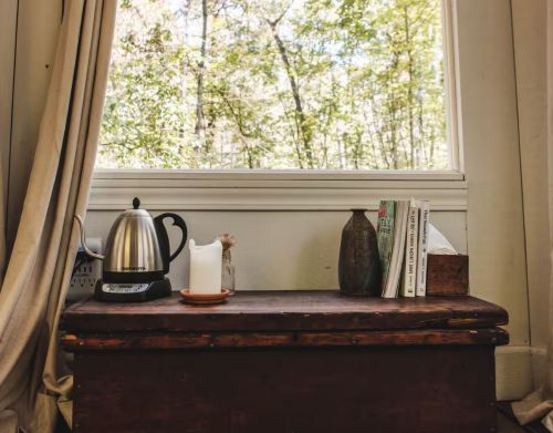 A kettle, candle, and decor on top of a wooden bench underneath a window.