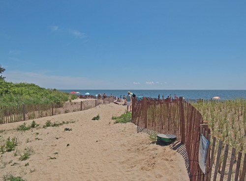 A sandy path with wooden fences leads to the beach, with people relaxing by the ocean under a clear sky.