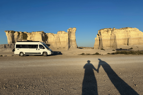 A camper van is parked in front of the chalk towers of Monument Rocks with the shadows of a couple holding hands seen on the ground.