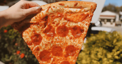 Hand holds a giant piece of pepperoni pizza outside.