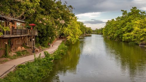 A darker river flows slowly next to a sidewalk and buildings with trees and flowers in Medicine Park, OK.