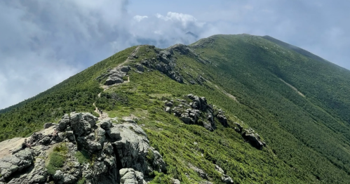 A narrow, winding hiking path follows a rocky, green alpine ridge under a partly cloudy sky on the Franconia Ridge Trail.