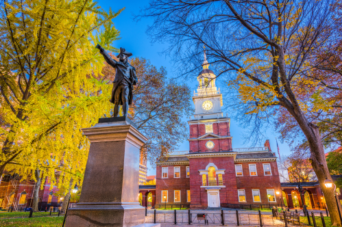 An illuminated statue of a Revolutionary War figure and Independence Hall's clock tower at dusk in Philadelphia.