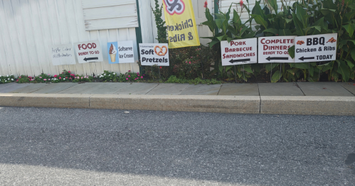 A collection of signs at the side of a house, each one labeled with a food item like "pork barbecue sandwiches" and "softserve ice cream"