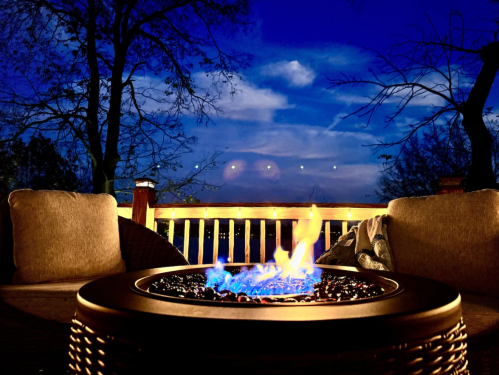 A lit fire pit with orange and blue flames flanked by two brown deck chairs in the foreground and a dark sunset in the background.