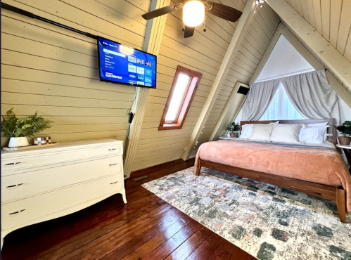 A bedroom at the top of the A-frame cabin with a bed, television mounted on the wall, and a white dresser. There is also a pink duvet on the bed.