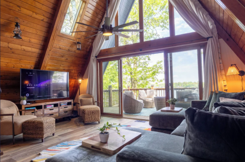 The cabin living room with huge picture wendows facing the lake and the back porch, a dark grey couch in the foreground, brown chairs and a flat screen TV in the background.