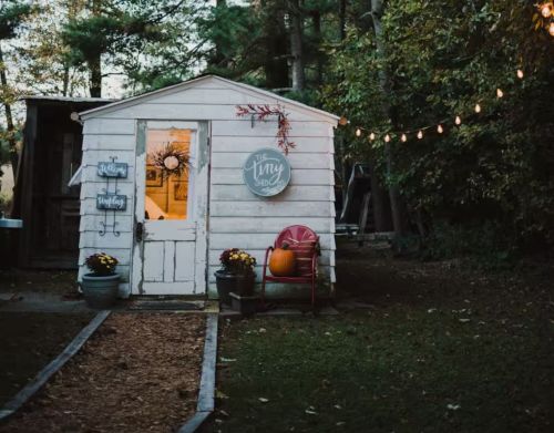 A small white shed that is serving as a tiny home with fall decorations on the outside.