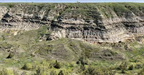 Green grass rising up to a brown rock face with a sliver of blue sky on the top