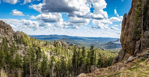view from atop a scenic highway with large forest trees, mountains, and a cloudy skies above in south dakota