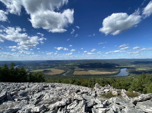 A wide expanse of blue sky dotted with clouds sits over a rocky outcrop that overlooks a flat valley of farmland and forests.