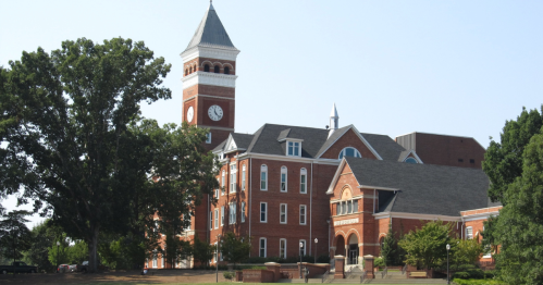 The historic Tillman Hall at Clemson, a red-brick building with a prominent clock tower, and a large tree on a green lawn.