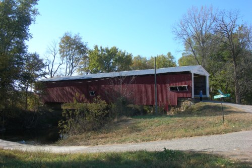 A red covered bridge leaning slightly, surrounded by green trees and grass, with a road sign nearby. Photo source: https://flic.kr/p/8LW9Qt