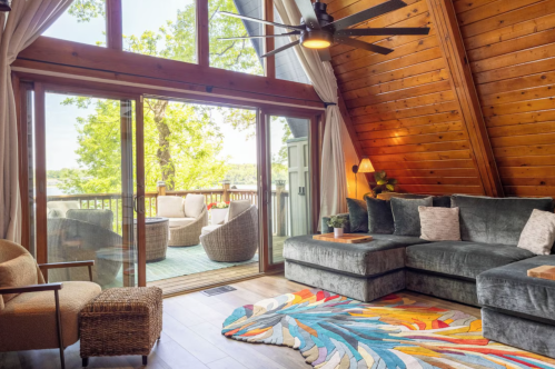 A living room in the cozy cabin featuring warm woods, a multi-colored rug, long grey couch and large windows.