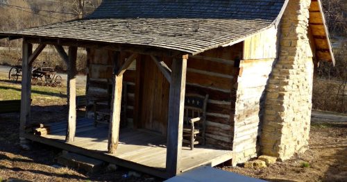 The historic brown, Didier Cabin in Brownsville, NE. It is a log cabin with a stone chimney on the right side and a brown wooden porch on the front with a dark colored roof.