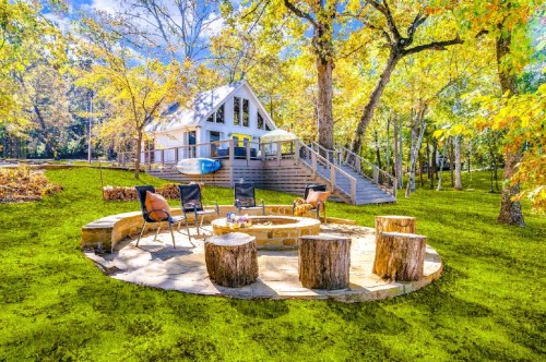 A large fire pit with a few chairs and wood stumps, tucked inside a yard with several golden trees, with a rustic cabin in the background at an Airbnb in Texas.