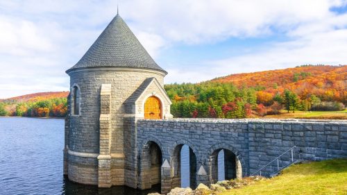 Views of the Saville Dam, one of the most scenic spots in CT, surrounded by a tranquil lake and lush forests with glowing autumn leaves.