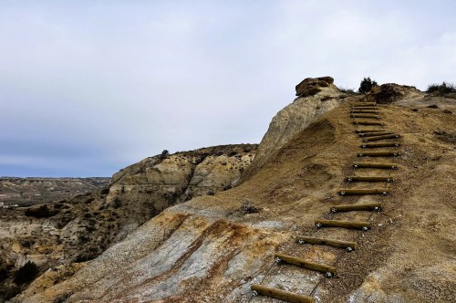 Views of wooden steps built into the side of a steep, rocky hill on an adventurous hiking trail in North Dakota, surrounded by a rugged landscape in the background.