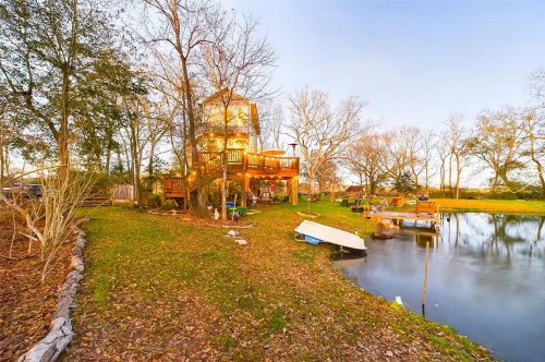A treehouse in Louisiana on the banks of a tranquil pond with a small dock and a canoe on the shore, surrounded by trees and a grassy yard with fallen leaves.