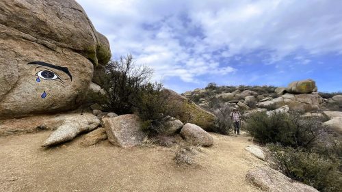 A lone hiker along a whimsical hiking trail in New Mexico, featuring a massive rock formation with an eye shedding tears painted on it, surrounded by other large rocks and desert shrubs.