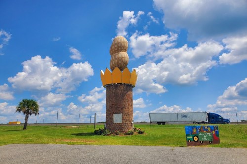 View of the world’s largest peanut, a towering monument in Georgia topped with a massive peanut and a golden crown, featuring a grassy lawn, a photo op sign, and vehicles along the highway in the background.
