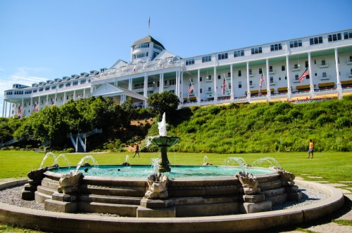 A grey stone fountain in the foreground with a lawn of bright green grass and the bright white Grand Hotel extending across the background