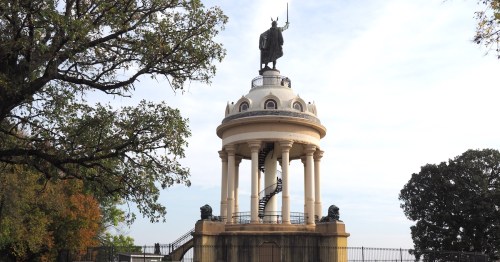A bronze, patinaed sculpture of a German warrior with a winged helmet stands atop a white iron cupola with a spiral staircase while holding a sword aloft.