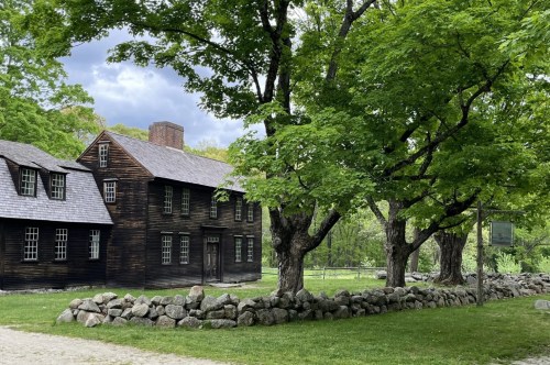 Alongside a path sits a historical wood house with a stone fence, green trees, and manicured lawn.