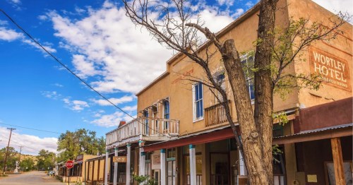 View of the abandoned Wortley Hotel located along the Turquoise Trail in Cerrillos, New Mexico, surrounded by other storefronts, an unpaved street, and a bare tree.