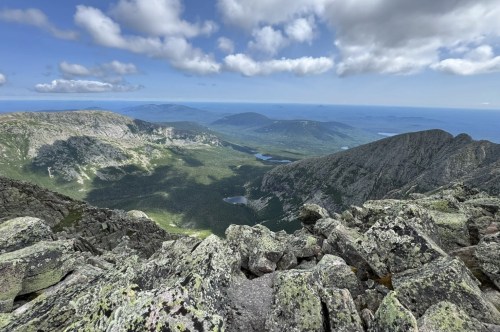 Sparse clouds hang over a large grouping of mountains, with forests and lakes nestled below.