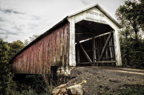 A faded, red, covered bridge spans a calm river, surrounded by greenery and under a partly cloudy sky. Image source: https://flic.kr/p/pPWaXX