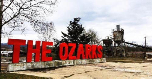 Large red sign reading 'THE OZARKS' in an outdoor setting, in Clinton, a rural town in Arkansas, with a factory in the background.