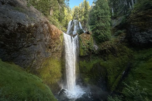 A two-part waterfall cascades down with mossy cliffs on either side and a forest surrounding it.
