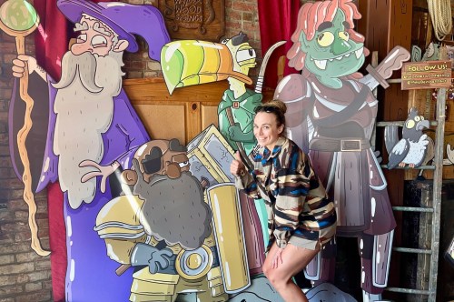 Four large cartoon figures are placed next to a brick wall as a woman in a tan jacket poses in front of them.