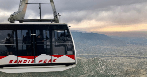 The Sandia Peak Tramway zooms across the frame, with a sunset sky against the mountains in the background