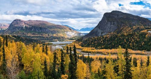 A scenic valley in autumn with vibrant yellow and orange foliage, a winding river, dense evergreen trees, and rugged mountains under a partly cloudy sky.