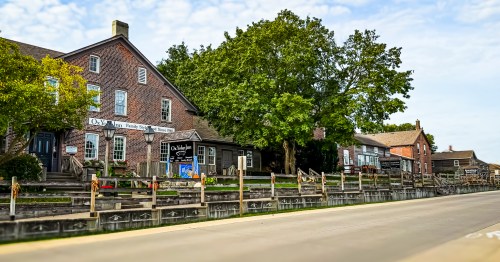 Row of brick buildings, vibrant green trees, and a sidewalk with a wooden fence on Main Steet Amama