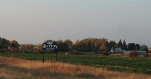 An Ashton, Idaho, town sign is in a field with the town and fall foliage behind it.