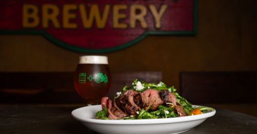 A flank steak in a salad with a beer behind it in front of a sign that says “Broadway”
