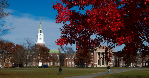 Red autumn leaves frame Dartmouth College's campus green with the clock tower and historic buildings under a blue sky.