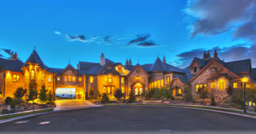 Castle-style mansion illuminated at dusk with stone exterior, turrets, and glowing windows.